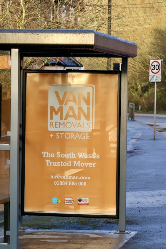 A photo of a bus stop by the side of the road with a Van Man Removals and Storage advertising sign